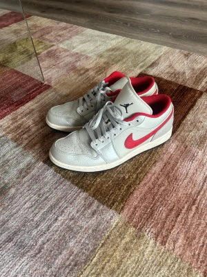 Nike Air Jordan 1 Low Night Track - Säljer ett par Nike Air Jordan 1 Low sneakers i metallic läder med röda detaljer och swoosh. Skorna har perforerad tå, grå snörning och röd insida. Klassisk Jumpman-logga på plösen och stilren siluett som passar till det mesta.