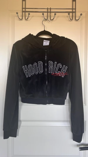 Svart velour hoodie från Hoodrich - Säljer en svart hoodie i mjuk velour från Hoodrich. Hoodien har dragkedja framtill, huva och broderad text 'HOODRICH' i vitt och rött på bröstet. Croppad modell med långa ärmar och bred mudd nedtill. Perfekt för en chill och trendig look.