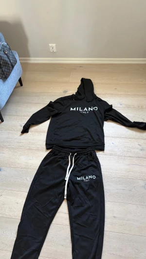 Svart Milano Italy set med huva - Säljer ett svart set med hoodie och byxor från Milano Italy. Hoodien har huva, magficka och vit text framtill. Byxorna har resår i midjan med vit dragsko och matchande vit text på benet. Perfekt för en avslappnad och trendig look.
