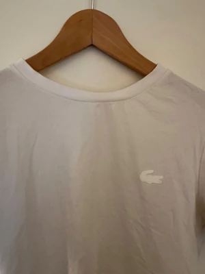 Vit t-shirt Lacoste  - Vit t-shirt med diskret broderad Lacoste logga i vitt. Baksidan har en dekorativ söm som går vertikalt längs ryggen. Liten i storlek. Passar S och XS