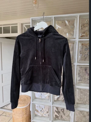 Svart velour hoodie Juicy Couture - Säljer en svart velour hoodie från Juicy Couture i storlek S. Hoodien har dragkedja, fickor framtill, justerbar huva med snören och diskret logga på bröstet. Perfekt för en chill och trendig look.