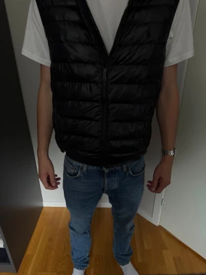 Svart dunväst med dragkedja - Snygg svart dunväst med quiltad design och dragkedja framtill. Västen har en klassisk passform och är perfekt att slänga över en hoodie eller t-shirt för extra värme. Tillverkad i glansigt material som ger en modern look.