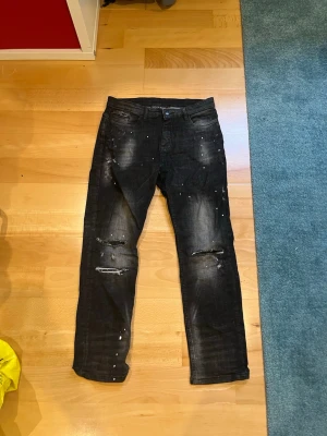 supply and demand jeans - Säljer ett par svarta jeans med slitningar och hål på båda benen samt vita färgstänk för en edgy look. Jeansen har fem fickor och normal passform. Perfekta för dig som gillar streetwear och vill ha en cool, avslappnad stil.