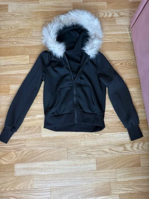 Svart hoodie med fuskpäls på huvan - Säljer en svart hoodie med dragkedja och stor huva kantad med vit fuskpäls. Hoodien har två fickor framtill och breda muddar vid ärmslut och midja. Perfekt för dig som vill ha en mysig och snygg look med lite extra attityd.
