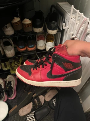Svart och röd Air Jordan 1 Mid - Säljer ett par Air Jordan 1 Mid sneakers i svart och rött läder med klassisk svart swoosh och röda detaljer. Skorna har svart snörning, vit mellansula och röd yttersula. Ikonisk Air Jordan-logga på sidan. Perfekta för dig som gillar streetwear och vill sticka ut.