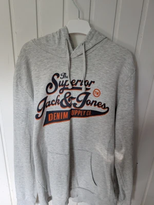 Grå hoodie Jack & Jones L - Snygg grå hoodie från Jack & Jones med trycket 'The Superior Jack & Jones Denim Supply Co' i marinblått och orange på bröstet. Klassisk huva med dragsko och stor magficka framtill. Mjuk insida och avslappnad passform, perfekt för chill dagar.