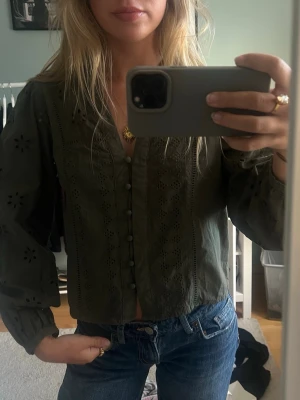 Mörkgrön blus med broderade detaljer - Snygg mörkgrön blus med broderade hålmönster och klädda knappar framtill. Blusen har långärmade puffärmar med resår vid ärmslut och en lätt v-ringad halsringning. Perfekt till jeans för en trendig look.