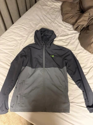 Under armour jacka - Snygg tvåfärgad vindjacka i svart och grått med hel dragkedja och huva. Jackan har två sidofickor och en liten grön logga på bröstet. Perfekt för blåsiga dagar och enkel att matcha med olika outfits.