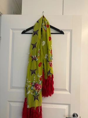 Grön sjal med broderi från Odd Molly - Snygg grön sjal från Odd Molly med broderade blommor och stjärnor i lila, rött och vitt. Sjalen har röda fransar i ändarna och är tillverkad i 65% bomull och 35% silke. Perfekt för att liva upp din outfit med färg och mönster.