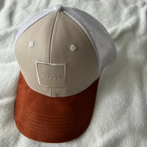 BOSS Beige och brun keps  - Snygg keps från BOSS med beige frontpanel och vit mesh baktill för extra ventilation. Skärmen är i en djup brun färg och har en mjuk, mockaliknande yta. Klassisk sexpanelsdesign med broderad BOSS-logga framtill.