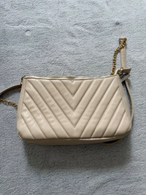 Beige väska med kedja - Snygg beige väska med V-mönster. Väskan har guldfärgad kedjedetalj på axelremmen och dragkedja upptill. Insidan är rymlig med en liten innerficka.