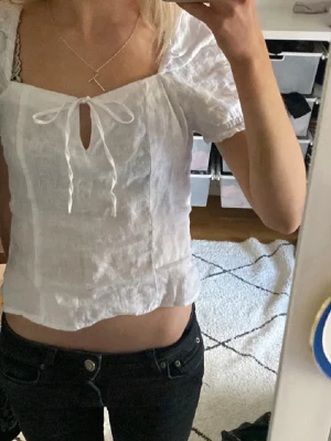 Vit blus med knyt - Söt vit blus i luftig bomull med korta färmar och knyt framtill. Modellen är croppad och har en lätt v-ringning. Perfekt för varma dagar och ger en romantisk vibe till din outfit.