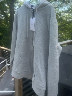 Grå zip hoodie från Moncler -  Med tags! Snygg grå zip hoodie från Moncler med huva och dragsko. Klassisk Moncler-logga på ärmen och praktiska fickor framtill. Tillverkad i mjukt material som känns skönt mot huden. Perfekt för en avslappnad och trendig look.