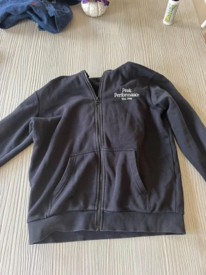 Svart hoodie från Peak Performance - Säljer en svart hoodie från Peak Performance med dragkedja framtill och två stora fickor. Tröjan har ribbade muddar vid ärmslut och nederkant samt logga broderad på bröstet. Tillverkad i mjukt material som känns skönt mot huden.