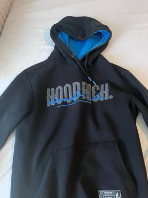 Svart hoodie från Hoodrich med blå detaljer - Snygg svart hoodie från Hoodrich med stor logga i grått och blått på bröstet samt blått foder i huvan. Har känguruficka framtill och tryck på ena ärmen. Perfekt för dig som gillar streetwear och vill sticka ut med coola detaljer.