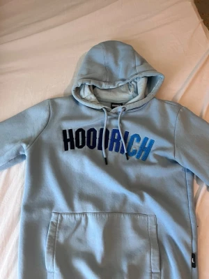 Ljusblå hoodie med HOODRICH-tryck - Ljusblå hoodie från Hoodrich med stor magficka och broderad text i svart och blått på bröstet. Klassisk huva med dragsko och mjukt material, perfekt för en chill och avslappnad stil. 