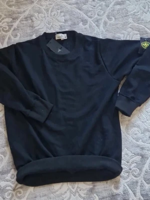 Svart sweatshirt från Stone Island - Säljer en svart sweatshirt från Stone Island med den klassiska loggan på vänster ärm. Tröjan har rund halsringning, ribbade muddar vid ärmslut och nederkant. Tillverkad i mjukt bomullsmaterial som är skönt att bära. Perfekt för en clean och stilren look.