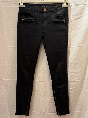 Svarta skinnjeans med dragkedjor - Snygga svarta skinnbyxot med guldfärgade dragkedjor vid fickorna och bensluten.Perfekta för en trendig och edgy look.