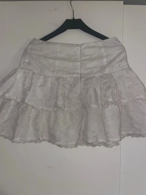 Vit volangkjol med broderade detaljer - Söt vit kortkjol med flera volanger och broderade blommönster. Kjolen har en feminin look med spetskant och är tillverkad i ett lätt bomullstyg som passar perfekt till varmare dagar. Dragkedja baktill för enkel påklädning.