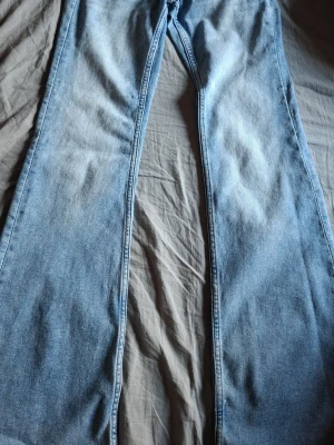 Ljusblå utsvängda jeans - Säljer ett par ljusblå, låga jeans med utsvängda ben och klassisk femficksdesign. Jeansen har hällor för skärp och stängs med knapp och dragkedja. Perfekta för dig som vill ha ett par bekväma jeans i denim.