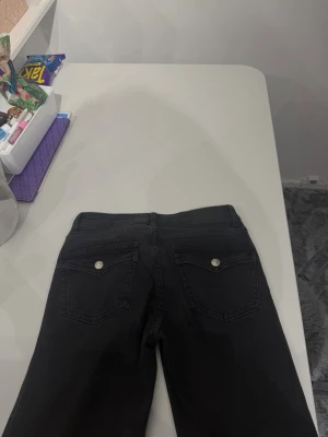 Svarta raka jeans (liten utsvängning) med fickor där bak  - Säljer ett par svarta jeans med raka ben och klassisk femficksmodell. Jeansen har silverfärgade knappar och detaljer både fram och bak. Materialet är denim med en mjuk känsla och de har låg midja. 