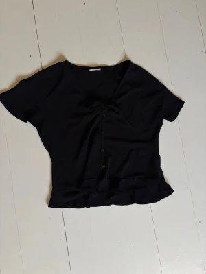 Svart blus med knappar från Vila - Svart blus från Vila med korta ärmar, v-ringning och knappar framtill. Blusen har en lätt rynkad detalj vid bysten och volangkant nedtill. Perfekt för en trendig och stilren look.