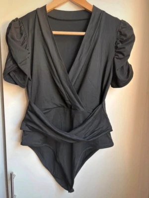 Svart omlott body med puffärm - Snygg svart body med omlott skärning och puffiga korta ärmar. Modellen har markerad midja och djup v-ringning framtill. Knappar i grenen. Aldrig använd!