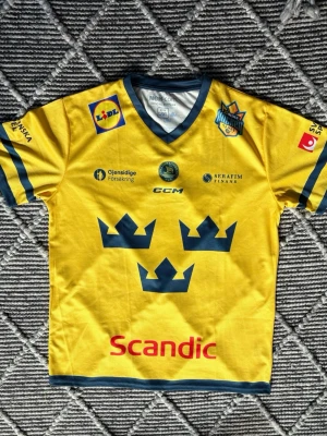 Gul Tre Kronor hockeytröja CCM - Säljer en gul Tre Kronor hockeytröja från CCM med blå detaljer och nummer 25 på ryggen. Tröjan har flera sponsorer tryckta på både fram- och baksida samt de klassiska tre kronorna på bröstet. Perfekt för dig som älskar svensk hockey.