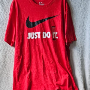 Röd Nike t-shirt med Just Do It-tryck - Riktigt snygg röd t-shirt från Nike med det klassiska svarta Swoosh-loggan och texten 'JUST DO IT.' i vitt framtill. Skön passform och tillverkad i mjukt bomullsmaterial. Perfekt för dig som gillar sportig och avslappnad stil.