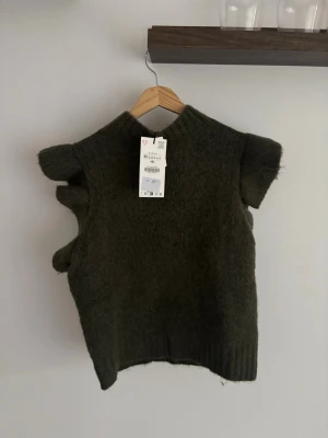 Mörkgrön stickad tröja med volang - Trendig mörkgrön stickad tröja med korta ärmar och volangdetaljer på sidorna. Västen har ribbstickad nederkant och är gjord i ett mjukt, lurvigt material som ger en mysig känsla. Perfekt att styla över en skjorta eller t-shirt för en cool lager-på-lager-look.