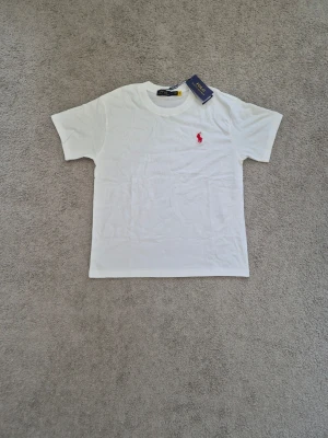 Polo Ralph Lauren - Klassisk vit t-shirt från Polo Ralph Lauren med röd broderad logga på bröstet. T-shirten har rund halsringning och korta ärmar. Tillverkad i mjuk bomull för en skön känsla och enkel stil.