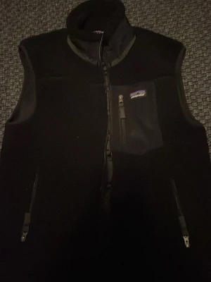 Svart fleeceväst från Patagonia - Svart fleeceväst från Patagonia med hög krage och hel dragkedja framtill. Västen har tre fickor med dragkedja, varav en bröstficka med logotyp. Perfekt för lager-på-lager och outdoor. Mjuk och varm känsla tack vare fleecematerialet. Använd väldigt få gånger ‼️‼️‼️