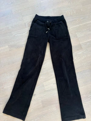 Svarta mjukisbyxor Juicy Couture XS - Säljer ett par svarta mjukisbyxor från Juicy Couture i storlek XS. Byxorna har raka ben, bred resår i midjan med dragsko och stora fickor framtill. Mjukt velourmaterial som är superbekvämt och trendigt.