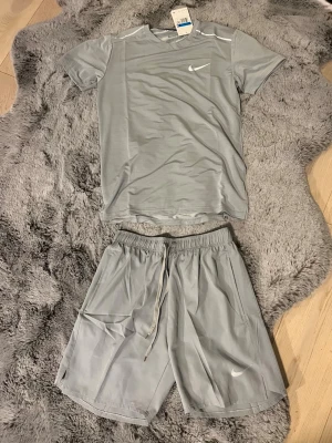 Grått träningsset från Nike - Snyggt grått träningsset från Nike med t-shirt och shorts. Båda plaggen har en minimalistisk design med Nike-logga på bröstet och benet. Tillverkade i ett lätt och snabbtorkande material som passar perfekt till träning och sportaktiviteter. Det står storlek M men passar S väldigt bra