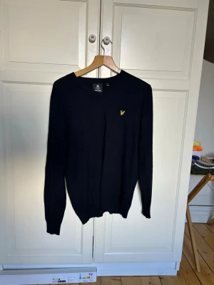 Svart stickad tröja från Lyle & Scott - Klassisk svart stickad tröja från Lyle & Scott med gul broderad logga på bröstet. Tröjan har V-ringning, långa ärmar och ribbade muddar. Perfekt till lager-på-lager och enkel att matcha med olika outfits.