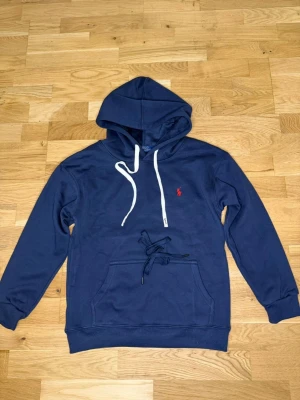 Mörkblå hoodie från Polo Ralph Lauren - Snygg mörkblå hoodie från Polo Ralph Lauren med klassisk röd logga broderad på bröstet. Hoodien har vit dragsko i huvan, känguruficka framtill och ribbade muddar. Perfekt för en chill och stilren look.