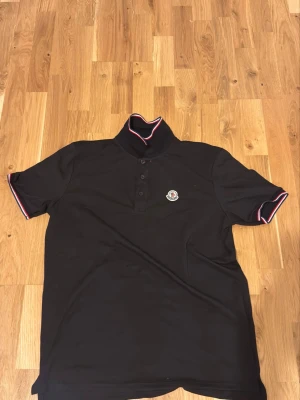 Svart Moncler pikétröja med krage - Svart pikétröja från Moncler med klassisk krage och knappslå framtill. Tröjan har korta ärmar med rödvita detaljer på ärmslut och krage samt Moncler-logga på bröstet. Perfekt för en stilren och sportig look. Storlek Xl men passar som L