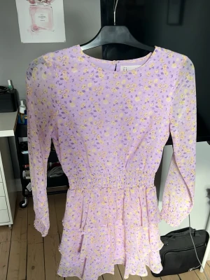 Ljuslila blommig volangklänning - Söt ljuslila kortklänning med små gula och lila blommor, långärmad och med volangdetaljer i kjolen. Klänningen har smock i midjan och vid ärmsluten för en bekväm passform. Perfekt för vår och sommar!