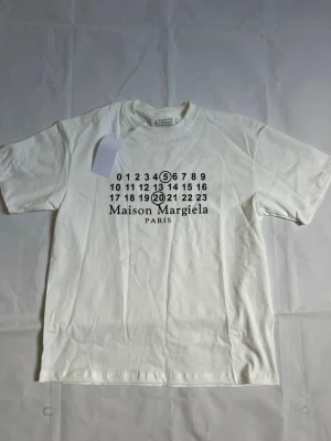 Vit Maison Margiela t-shirt - Vit t-shirt från Maison Margiela med svart tryck framtill som visar märkets ikoniska siffror och logga. Klassisk rund hals och korta ärmar. Tillverkad i mjuk bomull för en skön och avslappnad look.
