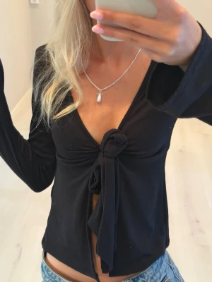 Topp med knyt - Trendig svart långärmad topp med djup v-ringning och knyt framtill. Toppen är croppad och har en smickrande passform som framhäver midjan. Perfekt att styla med jeans eller kjol för en cool look.