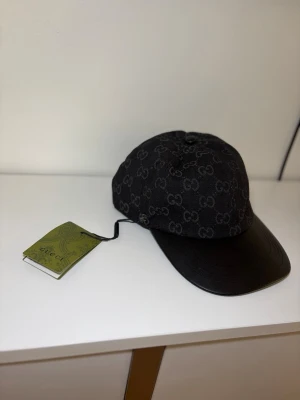 Svart keps med GG-mönster - Snygg svart keps med klassiskt GG-mönster över hela kepsen. Skärmen är i svart läder och resten av kepsen är i tyg. Perfekt accessoar för dig som vill ha en exklusiv och trendig look.