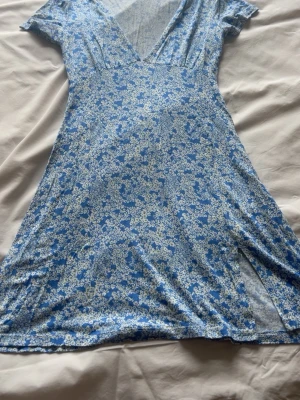 Blå blommig kortklänning med v-ringning - Söt blå kortklänning med vitt blommönster och v-ringad hals. Klänningen har korta ärmar och en lätt utställd kjol med slits. Perfekt för varma dagar och ger en fräsch sommarkänsla. Tillverkad i ett mjukt och följsamt material.