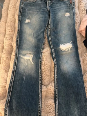 Blå slitna bootcut jeans Silver - Säljer ett par blå bootcut jeans från Silver med slitningar och hål på låren och knäna. Jeansen har klassisk femficksmodell, kontrastsömmar och låg midja. Perfekta för en avslappnad och trendig look.