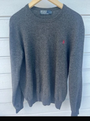 100 procent merinoull ullGrå stickad tröja från Polo Ralph Lauren - Mysig grå stickad tröja från Polo Ralph Lauren med rund halsringning och den klassiska röda logotypen broderad på bröstet. Tröjan har långa ärmar och ribbade muddar vid ärmslut och nederkant. Perfekt för lager på lager under kyliga dagar.