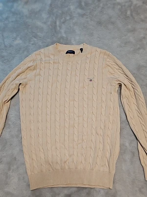 Beige kabelstickad tröja från Gant - Kabelstickad beige tröja från Gant med rund halsringning och diskret broderad logga på bröstet. Tröjan har långa ärmar och ribbade muddar vid ärmslut och nederkant. Perfekt för en stilren och klassisk look.