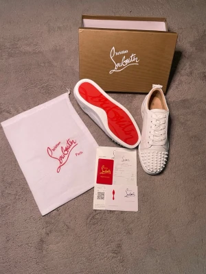 Vita Christian Louboutin sneakers - Snygga vita sneakers från Christian Louboutin med ikonisk röd sula och nitar på tån. Skorna har klassisk låg profil, vita skosnören och är tillverkade i läder. Perfekta för dig som vill sticka ut med exklusiv design och detaljer.