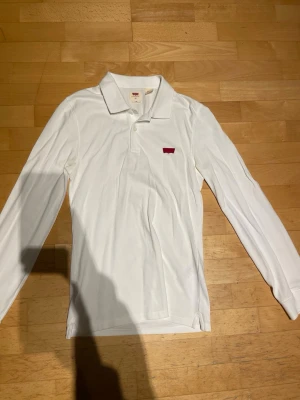 Vit långärmad skjorta från Levi's XS - Klassisk vit långärmad skjorta från Levi's i slim fit. Skjortan har krage, knappar framtill och den ikoniska röda Levi's-loggan på bröstet. Tillverkad i mjuk bomull som känns skön mot huden. Perfekt för en clean och stilren look.