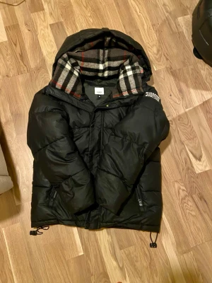 Svart pufferjacka från Burberry - Säljer en svart pufferjacka från Burberry med stor huva och klassiskt rutigt foder i grått, vitt, svart och rött. Jackan har dragkedja, tryckknappar och justerbara snören nedtill. Burberry-logga på vänster ärm. Perfekt för kalla vinterdagar.