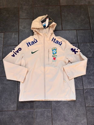 Beige Brasil Nike vindjacka med huva - Snygg beige vindjacka med huva från Nike, prydd med Brasilien fotbollslandslagets emblem och flera sponsorloggor i blått. Jackan har dragkedja framtill, två sidofickor och justerbara ärmslut. Perfekt för dig som gillar sportig stil och vill sticka ut.