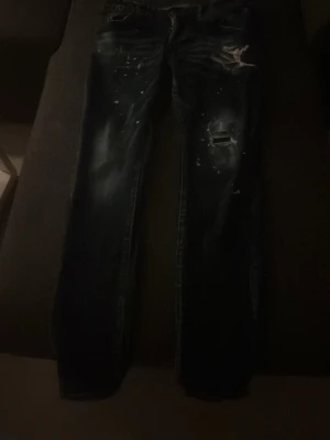 Mörkblå slitna jeans med hål - Säljer ett par mörkblå jeans med slitningar och hål på låret och knät. Jeansen har smal passform och coola detaljer med färgstänk och slitna partier. Perfekta för en avslappnad och trendig look.